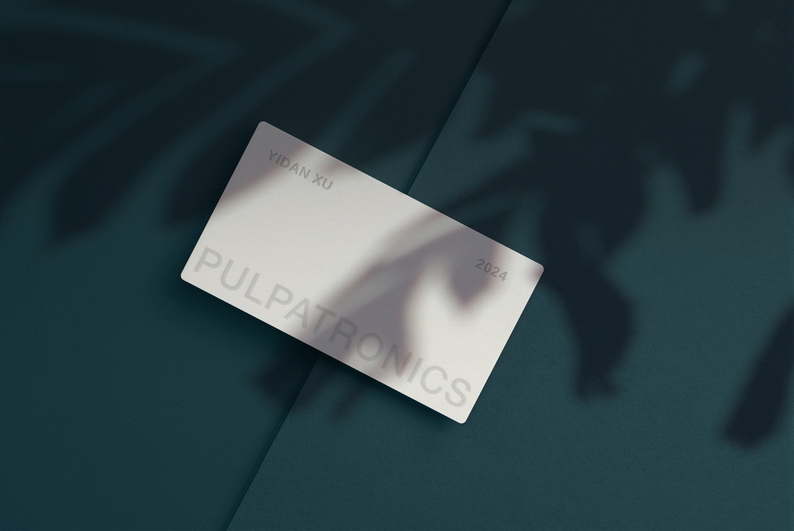 A PulpaTronics business card mockup: a paper-white card reading 'YIDAN XU', 'PULPATRONICS' and '2024' in embossed type, photographed on a deep teal surface under leaf shadows