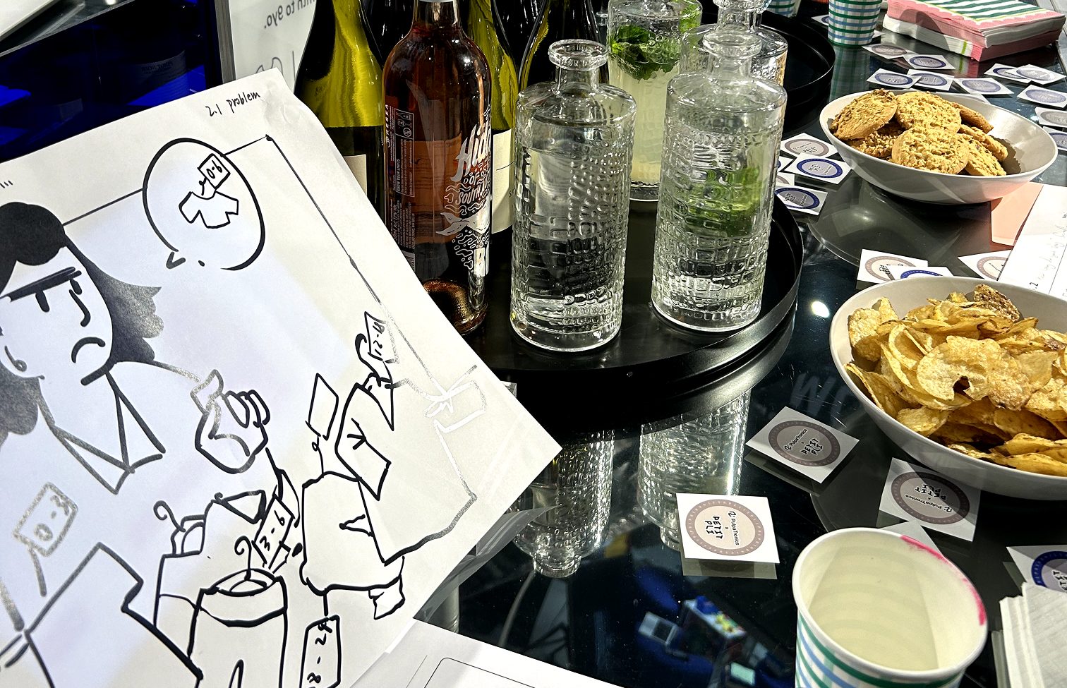A customer workshop table: printed illustration worksheets numbered by problem, branded PulpaTronics × Petit Pli stickers, glass carafes, crisps and biscuits