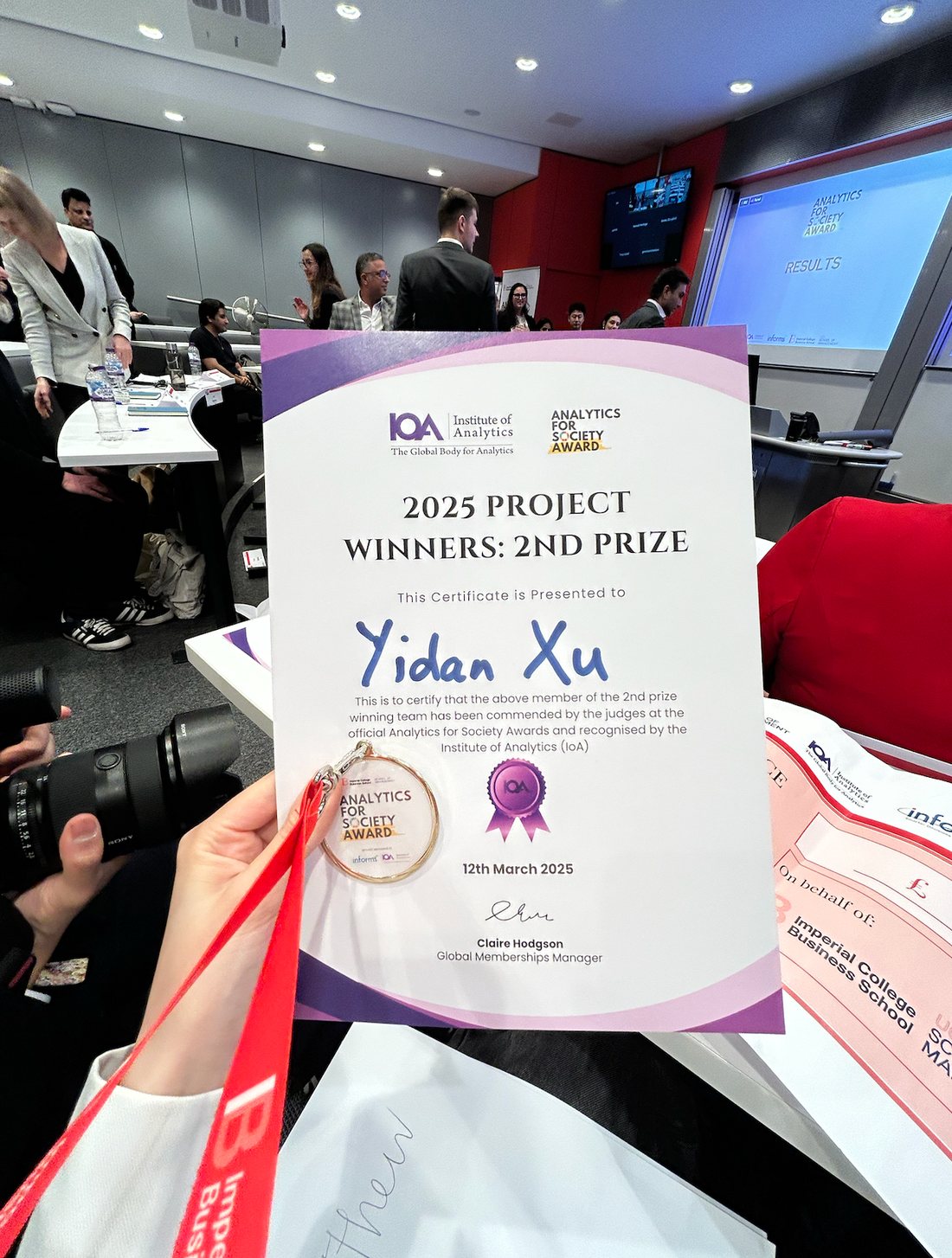 The Analytics for Society Award 2025 certificate from the Institute of Analytics, reading '2025 Project Winners: 2nd Prize' and presented to Yidan Xu, dated 12th March 2025, held up at the awards ceremony