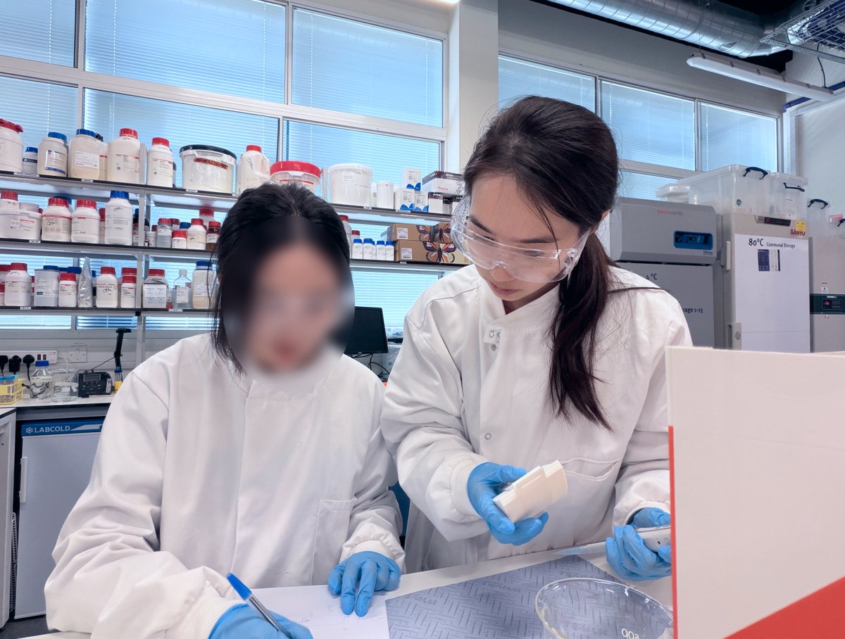 Yidan in a lab coat and safety glasses holding a white moulded tablet, working at a bench with a colleague whose face is blurred