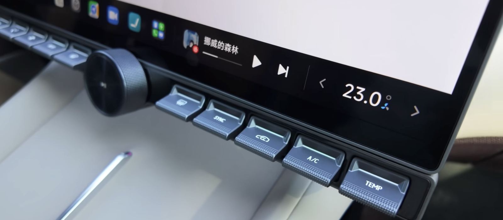 Close up of the physical key strip on the frame under the centre screen, with keys marked TEMP, A slash C, SYNC and demist and a knurled knob, and directly above them the bar on screen showing 23.0 degrees and the music controls